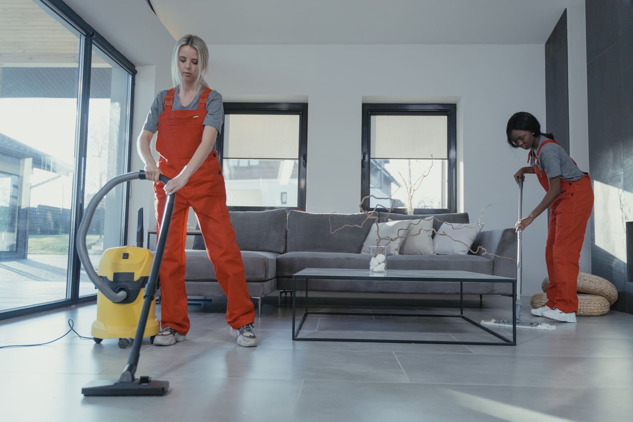 Two women wearing red overalls vacuuming and sweeping a contemporary living room.